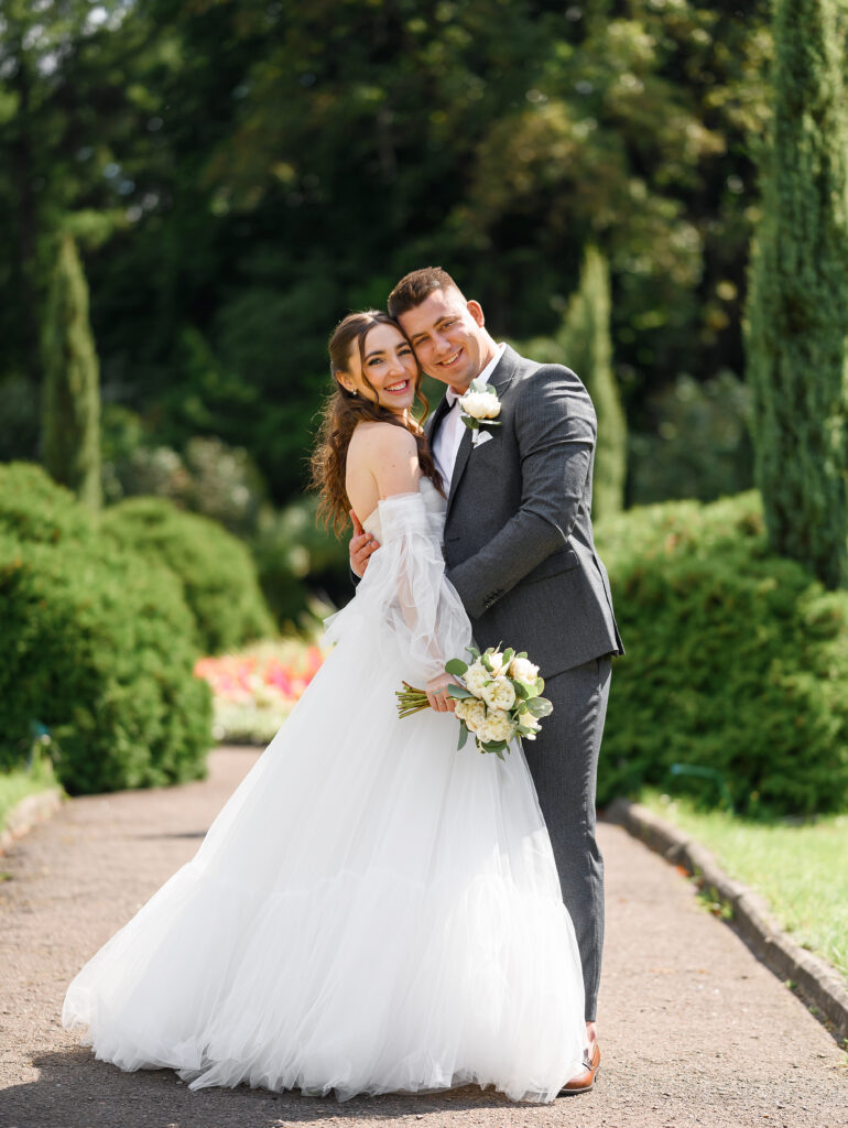 Portrait of happy married couple, handsome groom in stylish gray suit hugging bride in wedding dress with bouquet while posing in the garden. Wedding day ceremony. how to choose a wedding venue in Florida